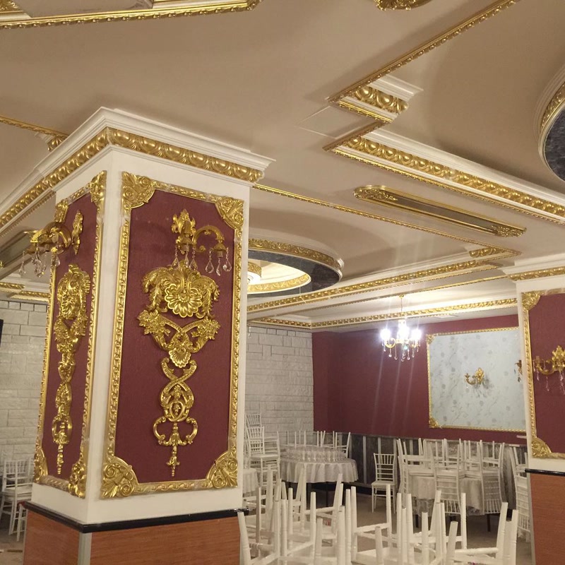 Poliuretan Profile și Elemente Decorative Clasic Sală Decorarea sălilor de nunți cu elemente din poliuretan de înaltă densitate (150-220 kg/m³ (țintă ~160 kg/m³)) este o soluție tehnică modernă pentru realizarea finisajelor interioare neoclasice de mare durabilitate. În această lucrare au fost utilizate profile cornișă P81142, ancadramente create cu șipci P82015 și colțare P82015C, alături de elemente ornamentale din seria P8088 (A, B, C, D, E) și panouri decorative P9135. Structura spumei poliuretanice rigide asigură o rezistență mecanică sporită și o absorbție de apă de sub 1%, prevenind fisurarea sau deformarea profilelor în spații mari supuse fluctuațiilor termice între -100 °C / +80 °C.

Montajul pe stâlpii masivi de susținere și pe suprafețele de tavan casetat s-a realizat prin lipire directă cu adeziv poliuretanic de structură, urmat de fixare mecanică temporară. Pentru obținerea îmbinărilor invizibile între profilele P81142 și șipcile P82015, rosturile au fost umplute cu mastic elastomeric special și șlefuite fin utilizând hârtie abrazivă cu granulație de 180-240. Timpul de maturare a adezivului de 24 de ore permite ajustarea precisă a colțarelor P82015C și alinierea impecabilă a reliefurilor pe fundalul de perete decorat.

Procesul de finisare a elementelor sculptate P8088 implică aplicarea unui grund acrilic de izolare, urmat de acoperirea cu vopsea pe bază de apă și patinare cu foiță sau vopsea aurie. Flexibilitatea profilului și stabilitatea dimensională a polimerului permit o aderență optimă a straturilor de vopsea fără risc de exfoliere. Integrarea sistemelor de iluminat ambiental și a aplicelor de perete pe panourile P9135 pune în valoare detaliile tridimensionale, oferind volumetrie și profunzime structurilor verticale fără a încărca greutatea proprie a pereților.

Pentru echipele de montaj și firmele de construcții, utilizarea elementelor presate din poliuretan reduce timpul de punere în operă cu peste 50% comparativ cu stucaturile clasice din ipsos. Greutatea redusă a elementelor ornamentale P8088 reduce sarcina structurală pe tavane și coloane, iar suprafața pretatată din fabrică garantează un consum redus de vopsea la fața locului. Această soluție tehnică asigură o durabilitate de lungă durată în spațiile comerciale cu trafic intens și cerințe ridicate de reprezentativitate.