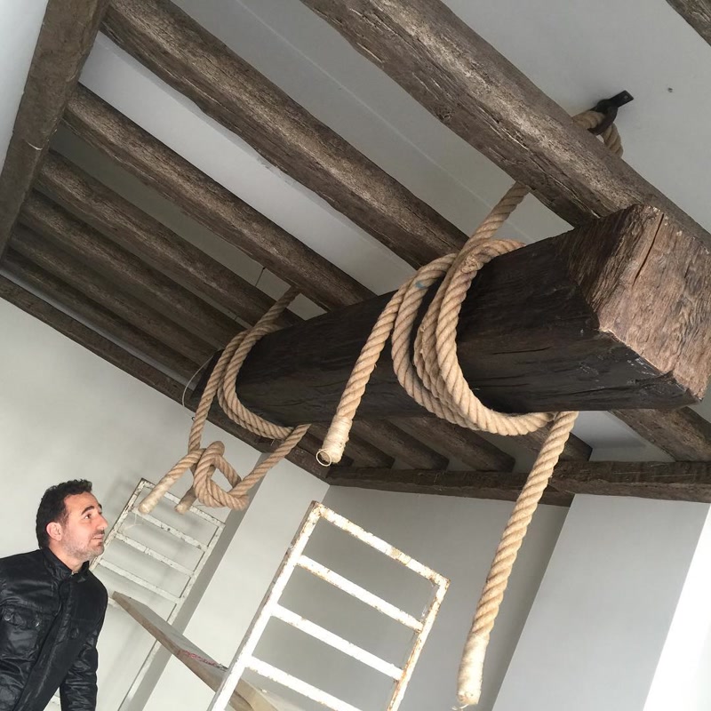 Grinzi Decorative din Poliuretan și Grindă Suspendată Grinzile decorative din poliuretan reprezintă elemente arhitecturale ușoare concepute pentru simularea structurilor masive din lemn pe tavanele spațiilor rezidențiale și comerciale. În această lucrare de amenajare rustică, tavanul alb servește drept suport pentru o rețea de căpriori (modele P1981B, P1981C și P1981D) și o grindă centrală suspendată prin intermediul unor frânghii naturale decorative. Utilizarea spumei poliuretanice de înaltă densitate, de aproximativ 150-220 kg/m³ (țintă ~160 kg/m³), permite obținerea unei texturi hiperrealiste de lemn cioplit, eliminând complet încărcarea structurală mare asupra planșeului.

Tehnica de montaj pentru sistemul de grinzi din poliuretan se bazează pe fixarea prealabilă pe tavan a unor șipci din lemn sau profile de ghidaj, peste care elementele decorative în formă de U se fixează prin lipire adezivă și ancorare mecanică discretă. Profilul principal P1981D are un interior gol ce permite mascarea traseelor electrice sau a canalelor tehnice, având o greutate redusă de doar 2-4 kg pe metru liniar, comparativ cu peste 80 kg pe metru în cazul unei grinzi din stejar masiv. Frânghiile montate prin împletire creează iluzia unei structuri suspendate, în timp ce susținerea reală este garantată de elementele de fixare ascunse.

Comportamentul poliuretanului în spațiile interioare este superior lemnului netratat în privința stabilității dimensionale, având o rată de absorbție a apei de sub 1% și o rezistență la temperaturi cuprinse între -100 °C / +80 °C. Materialul nu crapă, nu se deformează sub influența variațiilor de umiditate și este complet imun la atacul insectelor xilofage sau al mucegaiului. Suprafața modelelor P1981B și P1981C este pregătită din fabrică pentru vopsire, permițând aplicarea lacurilor sau a vopselelor acrilice pe bază de apă pentru obținerea nuanței exacte de nuc sau stejar închis.

Pentru echipele de montaj și firmele de construcții, utilizarea acestor grinzi din poliuretan reduce timpul de execuție pe șantier cu peste 70% față de manipularea și debitarea grinzilor din lemn uscat. Finisarea îmbinărilor se realizează simplu prin șlefuire ușoară cu hârtie abrazivă de granulație 180-240 și sigilare cu mastic poliuretanic adaptat. Sistemul decorativ astfel instalat oferă o durabilitate ridicată în timp fără a necesita tratamente periodice antifungice sau ignifuge costisitoare.