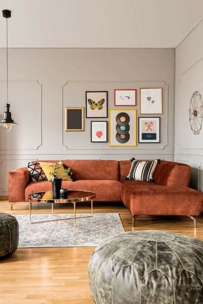 Aplicarea Baghetelor din Poliuretan P85002 pe Perete Accent Profilul din poliuretan P85002 este o baghetă decorativă liniară proiectată pentru realizarea casetamentelor pe pereții interiori în proiectele moderne și clasice. În amenajarea din imagine, montajul a fost efectuat direct pe suprafețe tencuite și amorsate, utilizând bagheta P85002 alături de colțarele speciale P85002M pentru îmbinări la 90 de grade fără tăieturi în unghi de 45 de grade. Spuma poliuretanică de înaltă densitate, având o masă volumică cuprinsă între 150 și 150-220 kg/m³ (țintă ~160 kg/m³), garantează o stabilitate dimensională excelentă și o rezistență sporită la impactul mecanic accidental. Spre deosebire de ipsosul tradițional, materialul nu crapă în timp și are o rată de absorbție a apei de sub 1%, ceea ce împiedică deformarea în condiții de umiditate fluctuantă.

Tehnologia de fixare a profilelor P85002 pe perete implică utilizarea unui adeziv poliuretanic structural cu aderență ridicată, aplicat continuu pe spatele profilului. Pentru alinierea perfectă a casetelor decorative, se trasează un caroiaj cu nivelă laser la o distanță de 15 cm față de plintă și cadrul ușilor. Utilizarea colțarelor prefabricate P85002M accelerează ritmul de lucru pe șantier cu peste 30%, eliminând erorile de debitare la îmbinările de colț. După lipire, rosturile de îmbinare dintre baghetă și colțar se umplu cu un mastic polimeric elastic, urmat de o șlefuire fină cu hârtie abrazivă de granulație 240 pentru obținerea unei suprafețe perfect omogene.

Suprafața baghetelor din poliuretan P85002 este acoperită din fabrică cu un grund alb pe bază de apă, pregătit pentru aplicarea vopselelor lavabile acrilice, poliuretanice sau latex. Polimerul își menține proprietățile mecanice și fizice pe un interval termic extins, cuprins între -100 °C / +80 °C, fiind ideal atât pentru spații rezidențiale încălzite, cât și pentru spații comerciale cu trafic intens. În comparație cu profilele din MDF sau lemn, poliuretanul nu suferă contracții la modificările de temperatură și nu este atacat de dăunători, asigurând o durabilitate de lungă durată fără necesitatea unor reparații periodice.

Structura casetată creată pe peretele principal servește drept cadru vizual pentru galeria de tablouri, evidențiind zona de relaxare definită de canapeaua modulară. Grosimea redusă a profilului P85002 permite integrarea discretă în planul peretelui, adăugând adâncime fără a reduce din spațiul util al încăperii. Proiectele de amenajare care folosesc elemente din poliuretan beneficiază de un timp de execuție redus și de costuri de întreținere minime, reprezentând o soluție tehnică eficientă pentru meșteri și firme de renovare.