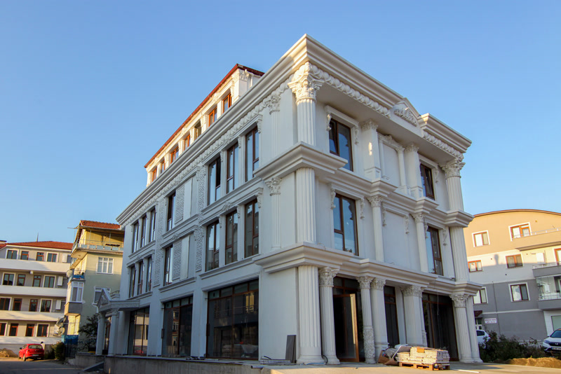 Poliuretan Profil Fațadă Clasică și Coloane Decor Profilul din poliuretan de înaltă densitate pentru fațade exterioare este un material compozit rigid cu o densitate structurală cuprinsă între 180 și 150-220 kg/m³ (țintă ~160 kg/m³), utilizat pentru placarea și decorarea arhitecturală a clădirilor cu mai multe niveluri. În realizarea acestei fațade neoclasice au fost integrate coloane structurale P5830, P5840 și P5860 alături de pilatri P5040 și P5040B, oferind o rezistență excelentă la ciclurile de îngheț-dezgheț în intervalul termic de la -100 °C / +80 °C. Spre deosebire de betonul arhitectural greu sau de polistirenul expandat vulnerabil la impact, poliuretanul își menține integritatea dimensională fără a suprasolicita structura de rezistență a clădirii.
Montajul profilelor de ancadrament P1456, P1459 și P1460 în jurul tâmplăriei exterioare se execută prin combinarea ancorării mecanice cu fixarea chimică. Pentru asigurarea unei etanșări hidrofuge complete, rosturile dintre elemente se tratează cu un adeziv poliuretanic special de îmbinare, lăsând un rost tehnologic de dilatare de minim 2 mm la fiecare racord structural. Panourile murale P8141 și consolele de susținere P9925 sunt fixate direct pe stratul de izolație termică sau zidărie folosind șuruburi din oțel inoxidabil și dibluri de ancorare structurală, prevenind formarea punților termice.
Performanța pe termen lung a suprafețelor din poliuretan este garantată de o absorbție de apă de sub 0.5%, ceea ce împiedică infiltrarea umidității și apariția fisurilor capilare. După finalizarea montajului mecanic, întreaga suprafață a profilelor, inclusiv detaliile decorative P2839 de pe frontoane, este grunduită și vopsită în 2 straturi succesive cu vopsea lavabilă de exterior pe bază de rășini acrilice sau siliconice. Această acoperire protejează polimerul împotriva radiațiilor ultraviolete și asigură o finisare cromatică omogenă și durabilă în timp.
Pentru constructorii și firmele de renovare, utilizarea sistemelor din poliuretan reduce timpul de punere în operă cu peste 50% comparativ cu stucaturile tradiționale din ipsos sau mortar. Greutatea redusă a elementelor decorative permite manipularea facilă pe schelă fără echipamente de ridicat grele, garantând în același timp o precizie geometrică absolută a profilelor pe fațadă. Rezultatul final combină estetica clasică anostă a clădirilor de elită cu parametrii tehnici moderni de izolație și rezistență mecanică.