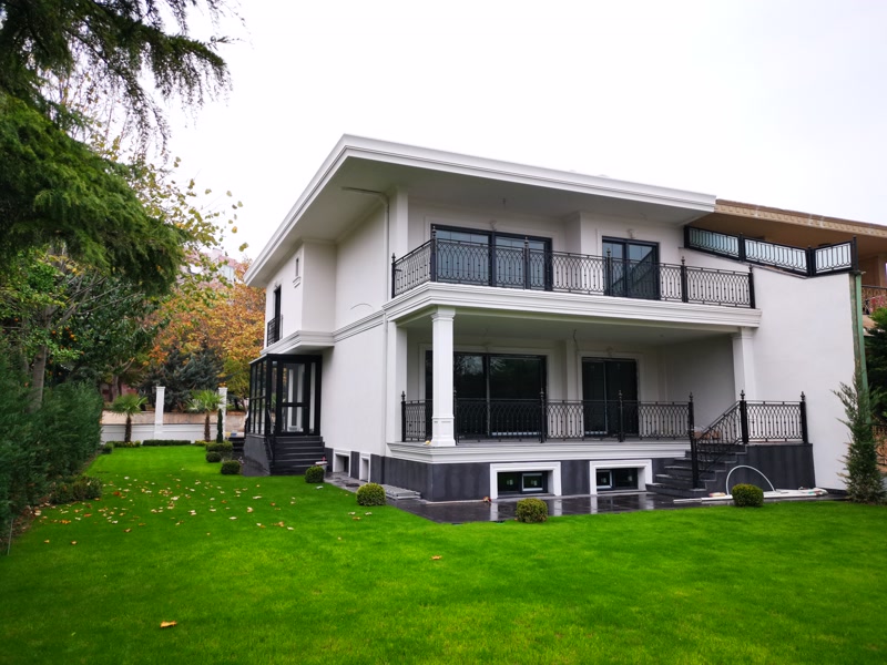 Sisteme din Poliuretan pentru Fațade de Vilă Modernă Profilul arhitectural din poliuretan dens pentru fațadă este un element decorativ și de protecție utilizat pentru conturarea ferestrelor, ușilor și a zonelor de tranziție pe suprafețele exterioare. În proiectul acestei vile moderne, s-au integrat profilele de ancadrament P1454, P1462, P1465 și P9001 pentru a crea rame volumetrice clare în jurul vitrajelor mari. În plus, pilatrii P5030 alături de bazele P5030B asigură o structurare verticală fermă pe terasa de la parter. Cu o densitate structurală cuprinsă între 180 și 150-220 kg/m³ (țintă ~160 kg/m³), aceste profile decorative oferă o rigiditate similară lemnului de esență tare, eliminând însă riscul de deformare în timp.

Tehnologia de instalare pe fațade izolate termic necesită respectarea riguroasă a etapelor de lipire și fixare mecanică. Profilele din poliuretan se montează direct pe stratul de masă de șpăcluire prin intermediul adezivilor poliuretanici monocomponenți, completați de dibluri de ancorare pentru elementele de mari dimensiuni. Materialul prezintă o rată de absorbție a apei de sub 0.5%, garantând o rezistență totală la îngheț și dezgheț în intervalul termic de la -100 °C / +80 °C. Îmbinările dintre profile se realizează cu adeziv special de sudură mecanică la îmbinări, prevenind apariția fisurilor capilare în zona de contact.

Comparativ cu soluțiile tradiționale din beton decorativ sau polistiren expandat (EPS), poliuretanul de înaltă densitate prezintă avantaje tehnice decisive pentru constructorii și aplicatorii. Spre deosebire de beton, greutatea redusă a poliuretanului nu încarcă structura de rezistență a clădirii și nu necesită ancoraje grele din oțel. Față de polistirenul simplu, suprafața poliuretanică este netedă, dură și rezistentă la șocuri mecanice accidentale, permisiunea de șlefuire fine asigurând o finisare impecabilă fără denivelări vizibile.

Finisajul final al ansamblului decorativ exterior se realizează prin aplicarea unei amorse speciale și a două straturi de vopsea lavabilă de exterior pe bază de rășini acrilice sau siliconice. Culorile deschise alese pentru această vilă sporesc reflexia luminii solare și protejează polimerul de radiațiile UV directe. Sistemul decorativ format din profilele P1454, P1462, P1465, P9001 și elementele P5030 completează arhitectura contemporană, oferind un aspect tehnic curat și o durabilitate de lungă durată pe întreaga suprafață a fațadei.