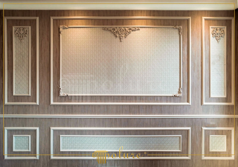 Panouri de Perete din Poliuretan și Profile Decorative Sistemul de casetare murală din poliuretan de înaltă densitate reprezintă o soluție tehnică eficientă pentru structurarea suprafețelor verticale în proiecte de amenajare interioară. Densitatea materialului de aproximativ 150-220 kg/m³ (țintă ~160 kg/m³) asigură o rezistență sporită la impact și stabilitate dimensională excelentă în intervalul de temperatură cuprins între -100 °C / +80 °C. Montajul acestor profile decorative permite mascarea imperfecțiunilor de planitate ale peretelui suport, oferind o bază rigidă și omogenă. Datorită absorbției de apă sub 0,1%, polimerul nu suferă modificări volumetrice, prevenind apariția fisurilor în zona îmbinărilor la variațiile de umiditate din încăpere.

Compoziția tehnică prezentată în această aplicație utilizează profile din poliuretan cu secțiuni specifice, precum P85002 pentru încadrarea principală și P85008 pentru detaliile interioare ale casetelor. Trecerea la nivelul de bază este marcată de un profil brâu P85016, stabilit pe linie orizontală continuă. Piese decorative speciale, precum elementele de colț P8081L și P8081R conectate cu piesa centrală P80111S, completează registrul superior prin fixare directă pe suprafața panoului. Pentru menținerea geometriei rectilinii pe unghiuri de 90 de grade, se utilizează piesa prefabricată P85002C, reducând timpul necesar debitărilor la 45 de grade în șantier.

Procesul de instalare profesională necesită pregătirea minuțioasă a suprafeței suport, care trebuie să fie curată, uscată și lipsită de praf. Fixarea profilului pe peretele cu finisaj texturat se realizat prin aplicarea unui adeziv structural pe bază de polimeri acrilici, completată de fixări temporare cu cuie de montaj. Pentru rosturile dintre baghete și colțare este obligatorie utilizarea unui adeziv de îmbinare poliuretanic special, aplicat uniform pe întreaga secțiune de contact. După întărirea completă a adezivului în decurs de 24 ore, excesul se îndepărtează prin răzuire mecanică, iar suprafața se finisează cu hârtie abrazivă de granulație P220 înainte de chituirea elastică.

Suprafața profilului din poliuretan este acoperită din fabrică cu un strat primar alb (amorsă acrilică), pregătit direct pentru vopsire. Finisajul final se execută prin aplicarea a două straturi de vopsea lavabilă, acrilică sau pe bază de apă, fără a fi necesară o șlefuire prealabilă intensivă. Comparativ cu elementele din ipsos tradițional sau lemn, poliuretanul nu crapă, nu absoarbe umiditatea și nu favorizează apariția mucegaiului. Această stabilitate mecanică și chimică face ca sistemul decorativ să fie ideal atât pentru spații rezidențiale, cât și pentru spații comerciale cu trafic mediu și ridicat.