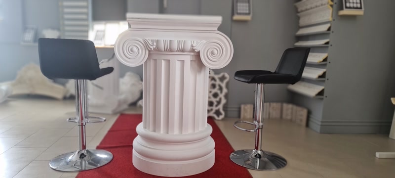 Stilp Decorativ din Poliuretan cu Capitel Ionic P5950B Coloana decorativă din poliuretan cu densitate înaltă reprezintă un element arhitectural structural-decorativ proiectat pentru finisaje interioare și exterioare de înaltă precizie. Sistemul modular prezentat în imagine este structurat din trei elemente principale: baza P5850, fusul vertical canelat P5250I și capitelul ionic ornamental P5950B. Fabricat din spumă poliuretanică densă de 150-220 kg/m³ (țintă ~160 kg/m³), acest ansamblu oferă o rezistență mecanică net superioară ipsosului clasic și un coeficient de absorpție a apei de sub 0.5%, eliminând riscul de igrasie sau degradare structurală în timp.

Tehnologia de instalare a coloanelor din poliuretan implică îmbinări adezive de înaltă rezistență și ancorare mecanică suplimentară unde este necesar. Fusul P5250I poate fi debitat cu ferăstraie mecanic cu dinți fini pentru a se potrivi exact la cota de înălțime a încăperii. Pentru lipirea componentelor P5850, P5250I și P5950B se utilizează adeziv poliuretanic structural pe bază de hibrid polimer, aplicat uniform pe fețele de contact. Timpul de priză al adezivului necesită 24 ore la o temperatură ambientală cuprinsă între +15°C și +25°C pentru o polimerizare optimă.

Etapele de finisaj tehnic presupun chituirea rosturilor de îmbinare cu un chit acrilic elastomeric, urmată de o șlefuire fină cu șmirghel de granulație 180-240. Suprafața elementelor P5950B și P5250I vine din fabrică acoperită cu un strat primar de amorsă albă, ceea ce permite vopsirea directă cu vopsele lavabile acrilice, poliuretanice sau alchidice fără conținut de solvenți agresivi. Materialul își păstrează stabilitatea dimensională pe o plajă termică extinsă de la -100 °C / +80 °C, fără a suferi dilatări sau contracții vizibile.

Pentru antreprenori și meșteri montatori, utilizarea profilatelor poliuretanice oferă un avantaj competitiv major reprezentat de viteza de punere în operă și greutatea redusă a materialului. O coloană completă asamblată din baze P5850, fus P5250I și capitel P5950B nu solicită încărcări suplimentare pe planșeu, putând fi manevrată fără echipamente grele de ridicat. În plus, profilul tubular interior permite îmbrăcarea facilă a stâlpilor metalici de rezistență sau a traseelor de instalații, combinând rolul de mascare tehnică cu cel de configurare spațială.
