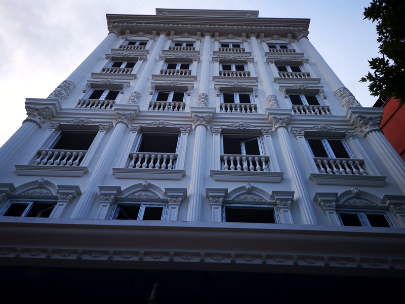 Ancadramente și Coloane din Poliuretan Fațadă Hotel Profilarea fațadelor clasice cu poliuretan de înaltă densitate reprezintă o soluție tehnică avansată pentru clădiri cu regim mare de înălțime, precum hotelul prezentat în proiect. Elementele arhitectural fabricate din poliuretan durabil, având o densitate medie cuprinsă între 150-220 kg/m³ (țintă ~160 kg/m³) și 150-220 kg/m³ (țintă ~160 kg/m³), oferă o rezistență mecanică sporită la factorii de mediu fără a încărca structura portantă a clădirii. Spre deosebire de betonul decorativ sau ipsosul tradițional, poliuretanul pentru fațadă elimină complet punțile termice datorită coeficientului scăzut de conductivitate termică. În acest proiect de hotel, ritmul vertical al fațadei este definit prin pilaștrii P5024A și sistemele de coloane P5540, P5560, P5840 și P5860, obținându-se o volumetrie simetrică și bine structurată.

Instalarea ancadramentelor de geam din gama P1460, P1460M40, P1462 și P1474 se realizează prin lipire structurală combinată cu ancorare mecanică invizibilă. Fiecare fereastră beneficiază de o conturare precisă, unde piesa superioară de fronton P2839 și arcele semicirculare P1456D160VIA / P1456D160VIB asigură o evacuare eficientă a apei pluviale, prevenind infiltrațiile la nivelul zidăriei. Datorită ratei de absorbție a apei de sub 0,1%, elementele decorative își păstrează stabilitatea dimensională pe o plajă largă de temperaturi extreme, de la -100 °C / +80 °C. Această rezistență hidrofugă previne fisurarea la îngheț-dezgheț, o problemă frecventă în cazul tencuielilor clasice sau a profilaturilor din polistiren netratat.

Sistemul de susținere vizuală este completat de consolele profilate P6003 montate la baza pervazurilor și sub cornișele principale, asigurând distribuția optică a încărcării. Procesul de montaj impune utilizarea adezivilor poliuretanici monocomponenți cu timp de procesare de 15 minute și întărire completă în 24 ore. Rosturile de îmbinare dintre profile se tratează cu adeziv special de sudură la rece pentru poliuretan, garantând monolitizarea suprafețelor și eliminarea posibilității de apariție a fisurilor micro-structurale. Finisajul exterior se realizează prin aplicarea a două straturi de vopsea elastomerică pe bază de apă, rezistentă la razele UV.

Utilizarea elementelor din poliuretan pe fațadele clădirilor comerciale reduce semnificativ costurile de întreținere pe termen lung și scurtează durata de execuție a lucrărilor cu până la 50% comparativ cu sistemele clasice din piatră sau ciment. Greutatea redusă a materialului simplifică manipularea pe schelă și elimină necesitatea structurilor auxiliare de sprijin extrem de grele. Totodată, suprafața amorsată din fabrică oferă o aderență excelentă pentru finisajele de fațadă, asigurând o durabilitate de peste 25 de ani în condiții optime de exploatare.