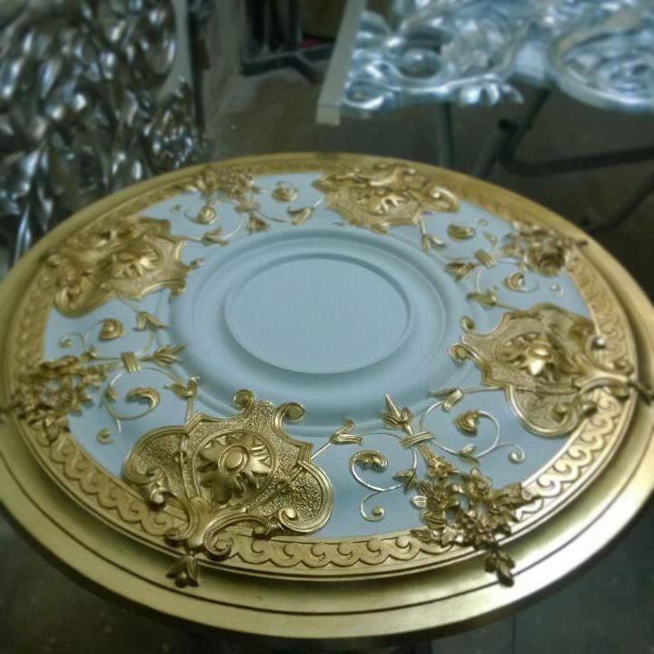 Rozetă din Poliuretan P721 pentru Decorarea Tavanului Rozeta din poliuretan P721 este un element decorativ circular conceput pentru structurarea estetică a tavanelor în proiecte rezidențiale de tip vilă sau spații comerciale clasice. Fabricată din spumă poliuretanică de înaltă densitate, având o valoare de aproximativ 150-220 kg/m³ (țintă ~160 kg/m³), această rozetă oferă o rezistență mecanică ridicată și o finisare detaliată a reliefurilor decorative. Spre deosebire de ipsosul tradițional, poliuretanul previne apariția fisurilor în timp și garantează o toleranță dimensională excelentă, facilitând o aliniere perfectă la nivelul suprafeței de montaj.

În faza de pregătire a suprafeței, piesa expusă prezintă o combinație cromatică între un fond albastru pastel mat și accente din vopsea acrilică sau patină aurie aplicată pe ornamentele în relief. Datorită structurii celulare închise a poliuretanului, rata de absorbție a apei este de sub 1%, ceea ce împiedică umflarea materialului la aplicarea vopselelor pe bază de apă. Pentru o aderență optimă a straturilor decorative, se recomandă șlefuirea ușoară a suprafeței primare cu hârtie abrazivă de granulație 240, urmată de aplicarea a cel puțin 2 straturi de finisaj protector.

Fixarea mecanică și structurală a rozetei P721 se realizată pe un tavan bine curățat și amorsat, utilizând adezivi polimerici sau poliuretanici cu priză rapidă. Datorită greutății reduse a piesei în comparație cu variantele din ipsos, nu este necesară ancorarea mecanică grea prin dibluri metalice, reducând riscul de deteriorare a structurii tavanului. Rezistența termică a poliuretanului, cuprinsă între -100 °C / +80 °C, permite instalarea rozetei în proximitatea corpurilor de iluminat de tip candelabru, fără risc de deformare la căldură emisă de sursa luminoasă.

Pentru firmele de construcții și echipele de finisaje interioare, rozeta P721 reprezintă o soluție eficientă din punct de vedere al timpului de executare a lucrărilor. Îmbinările perimetrale dintre rozetă și placa de gips-carton se sigilează cu un chit acrilic elastic, prevenind fisurile cauzate de dilatările structurale ale clădirii. Rezultatul final oferă o durabilitate crescută pe termen lung, eliminând necesitatea restaurărilor frecvente specifice profilaturilor clasice din ipsos.