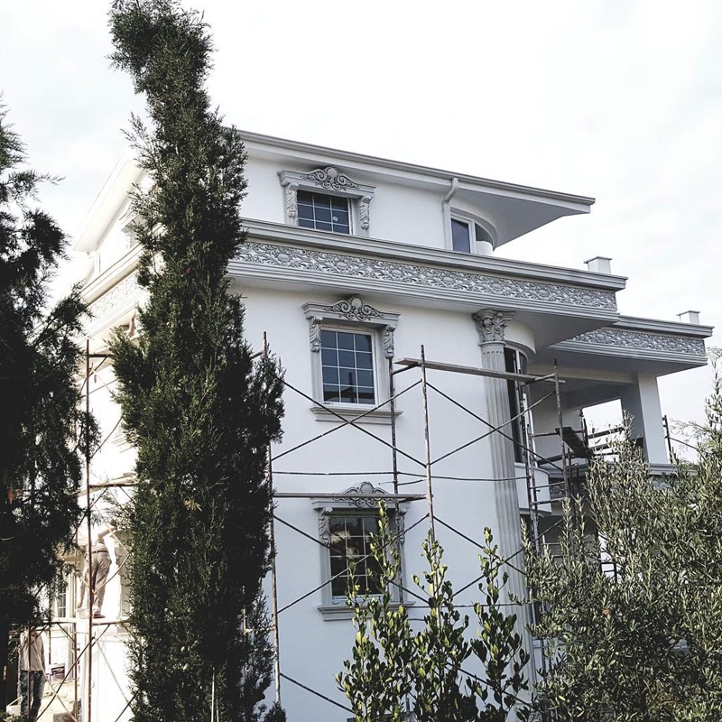 Poliuretan Profile Fatada si Ancadramente Geam Vila Poliuretanul de înaltă densitate pentru exterior este un material compozit rigid utilizat pentru finisarea arhitecturală a fațadelor rezidențiale, având o densitate medie de 150-220 kg/m³ (țintă ~160 kg/m³) și rezistență ridicată la intemperii. În acest proiect de vilă cu arhitectură clasică, montajul profilelor ornamentale se execută pe o structură termoizolantă existentă, unde este necesară o fixare precisă și durabilă. Sistemul folosește socluri și ancadramente verticale compuse din profilele P1454, P1456 și P1459 pentru realizarea contururilor rigide la ferestre. Spre deosebire de elementele tradiționale din beton sau EPS expandat, spuma poliuretanică densă nu absoarbe apa (absorbție sub 1%), prevenind astfel fisurarea în ciclurile îngheț-dezgheț și menținând integritatea structurală a îmbinărilor pe termen lung.

Tehnologia de aplicare presupune combinarea lipirii structurale cu ancorarea mecanică acolo unde sarcinile consolațiunii sunt mai mari. Frontoanele ornamentale P2839 și consolele de susținere P9925 și P9927 sunt fixate pe fațadă prin adeziv poliuretanic monocomponent de înaltă aderență și dibluri din oțel inoxidabil încastrate. Pentru zona de sub streașină și brâurile intermediare, s-au integrat panouri decorative cu relief adânc din seria P8107, care necesită o aliniere milimetrică la îmbinările de colț. Utilizarea coloanelor decorative P5840 adaugă stabilitate vizuală și susținere estetică balcoanelor, profilele având un comportament stabil la variații termice extreme cuprinse între -100 °C / +80 °C.

Etanșarea îmbinărilor între profilele din poliuretan se realizează utilizând adeziv special de îmbinare pe bază de poliuretan reacționar, urmată de șlefuirea fină cu hârtie abrazivă de granulație 220 după uscarea completă. Această etapă tehnică este critică pentru eliminarea punților termice și a microfisurilor de dilatare. Toate suprafețele din poliuretan vin pre-amorsate din fabrică cu un strat protector, fiind pregătite direct pentru aplicarea vopselelor lavabile de exterior pe bază de rășini acrilice sau siliconice. Calitatea suprafeței permite o acoperire omogenă și reflexii uniforme ale luminii, punând în evidență detaliile sculpturale fără a fi nevoie de straturi groase de tencuială.

Pentru firmele de construcții și montatori, utilizarea elementelor ornamentale din poliuretan reduce considerabil timpul de punere în operă și sarcina totală pe structura clădirii. Greutatea redusă a pieselor elimină necesitatea utilajelor grele de ridicare în timpul instalării pe schelă. Rezistența mecanică ridicată și stabilitatea dimensională asigură o mentenanță minimă în timp, fațada necesitând doar spălare periodică cu apă sub presiune joasă. Soluția integrată oferă un randament superior față de stucaturile clasice din ipsos sau ciment, garantând o finisare impecabilă fără risc de decojire sau degradare biologică.