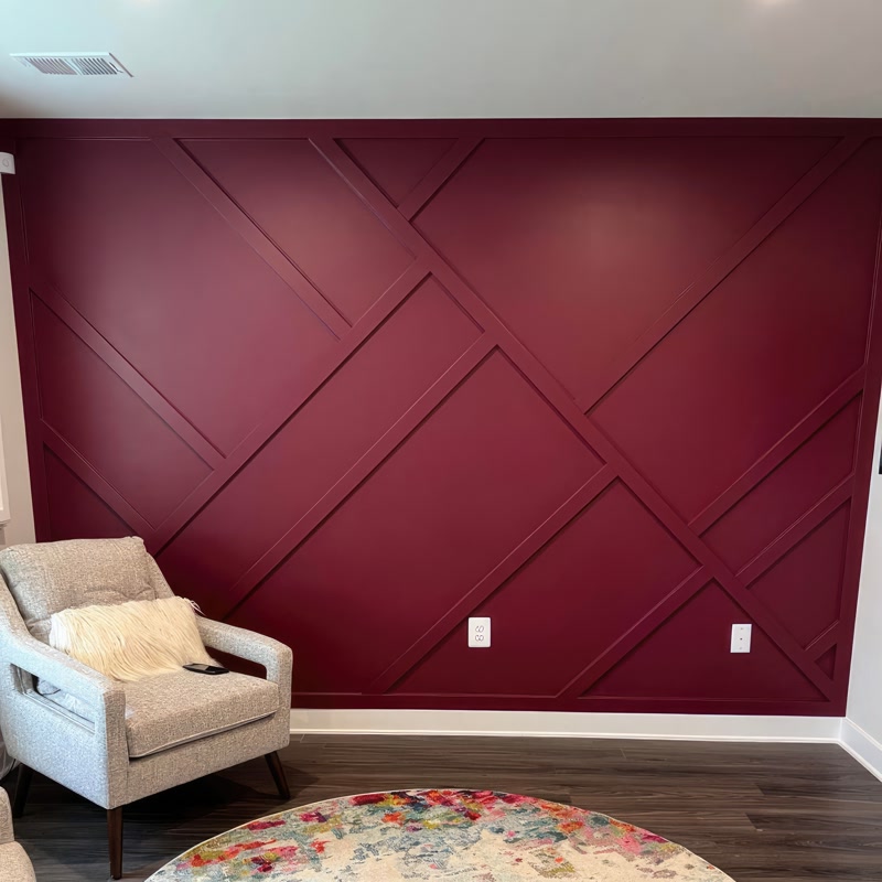 Poliuretan Geometric Decorativ pentru Perete Accent Vișiniu Aplicarea profilelor decorative din poliuretan P851350 pe un perete accent reprezintă o tehnică modernă de finisare interioară ce oferă dinamică vizuală spațiului prin linii geometrice asimetrice. Această lucrare tehnică se realizează prin fixarea profilelor poliuretanice de mare densitate (aproximativ 150-220 kg/m³ (țintă ~160 kg/m³)) direct pe suprafața gips-cartonului sau a tencuielii pregătite în prealabil. Structura rigidă și stabilitatea dimensională a poliuretanului împiedică deformarea în timp, având o rată de absorbție a apei de sub 1%, ceea ce garantează o aderență excelentă a vopselelor lavabile și o rezistență sporită în medii cu umiditate variabilă.

Procesul de montaj necesită o planificare riguroasă a caroiajului și executarea tăieturilor la unghi precis de 45 sau 90 de grade, în funcție de intersecțiile prevăzute în proiectul de design. Pentru fixarea profilelor P851350 se utilizează un adeziv de montaj pe bază de poliuretan sau polimer hibrid, aplicat în cordon continuu pe spatele elementului. La îmbinările dintre profile este obligatorie folosirea unui adeziv special pentru rosturi, urmată de o șlefuire fină cu hârtie abrazivă de granulație 240-320 după uscare, asigurând o suprafață perfect continuă înainte de vopsire.

Din punct de vedere al comportamentului materialului, poliuretanul prezintă avantaje tehnice net superioare elementelor clasice din lemn sau ipsos. Rezistența termică cuprinsă între -100 °C / +80 °C permite instalarea în apropierea corpurilor de încălzire fără risc de fisurare sau contracție. În plus, greutatea redusă a profilelor P851350 simplifică operarea pe șantier și reduce timpul de punere în operă cu până la 40% comparativ cu stucaturile tradiționale din ipsos, eliminând necesitatea sistemelor mecanice grele de ancorare.

Etapa de finisare implică aplicarea unui strat de grund primar, urmat de două straturi de vopsea acrilică sau lavabilă mată, precum nuanța de bordo intens prezentată în aplicația din imagine. Suprafața amorsată din fabrică a profilului P851350 optimizează consumul de vopsea și asigură o acoperire omogenă pe întreaga structură geometrică. Această abordare tehnică garantează o durabilitate de lungă durată și un finisaj tridimensional curat, ideal pentru proiecte rezidențiale de nivel înalt.