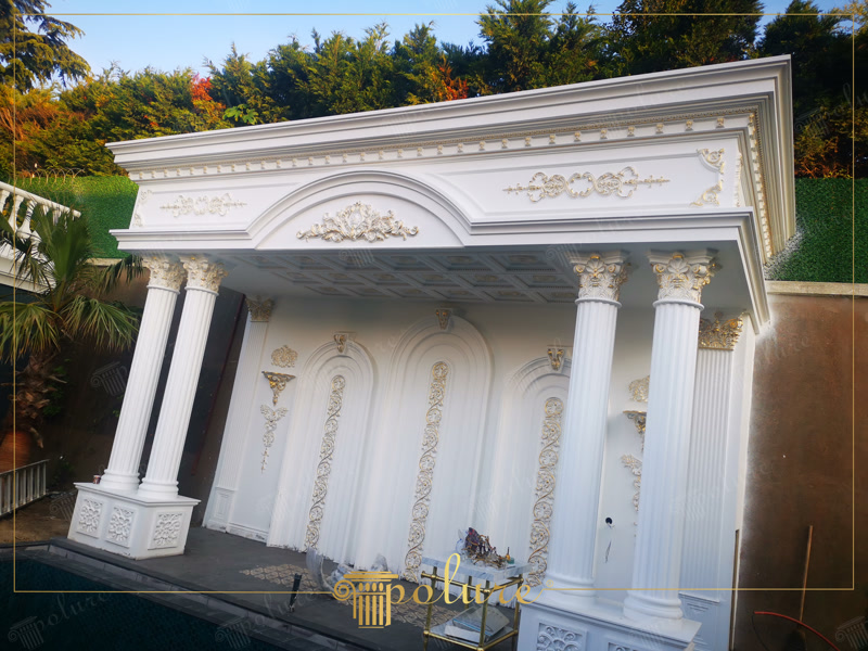 Pavilion Exterior Clasica cu Coloane si Profile Poliuretan Structura decorativă exterioară realizată din elemente din poliuretan de înaltă densitate reprezintă o soluție tehnică durabilă pentru finisarea arhitecturală a pavilioanelor de grădină și a foișoarelor de vilă. Cu o densitate structurală cuprinsă între 180 și 150-220 kg/m³ (țintă ~160 kg/m³), aceste profile poliuretanice nu absorb apa (absorbție sub 0,5%) și își mențin stabilitatea dimensională la variații extreme de temperatură între -100 °C / +80 °C. Spre deosebire de betonul amprentat sau ghipsul tradițional, materialul nu crapă în urma ciclurilor de îngheț-dezgheț, eliminând riscul de exfoliere a straturilor exterioare.

Montajul sistemului de coloane P5840 și al capitulurilor P5940B necesită un miez de susținere din oțel sau beton, poliuretanul acționând ca o teacă decorativă rezistentă la intemperii. Fixarea elementelor verticale se realizează prin ancorare mecanică combinată cu adezivi structurali poliuretanici monocomponenți. În zona de perete din spate, pilaștrii P5040 și P5040B sunt aplicați direct pe tencuială prin lipire în puncte și cordoane continue, asigurând o aliniere milimetrică și un transfer redus al tensiunilor structurale.

Realizarea arcelor semicirculare superioare și a ancadramentelor s-a executat prin îmbinarea profilelor flexibile P1456 împreună cu elementele de boltă P1456D160VIC și P1456D230VI. Rosturile de dilatație de 2-3 mm dintre profile sunt sigilate cu mastic elastomeric poliuretanic pentru a preveni fisurile cauzate de mișcările naturale ale clădirii. Detaliile ornamentale precum coronamentele P2833 și casetele de tavan P7308 sunt îmbinate prin debitare la unghi de 45 de grade și șlefuite ulterior cu hârtie abrazivă de granulație 180-240 înainte de vopsire.

Procesul de finisare a ansamblului exterior folosește amorse acrilice pe bază de apă și vopsele elastice rezistente la razele UV. Accentele decorative aplicate pe elementele P8088A și P507 sunt realizate cu vopsele metalizate pe bază de rășini sintetice, garantând o retenție excelentă a culorii în timp. Această tehnică de punere în operă garantează o durată de viață a fațadei de peste 25 de ani fără restaurări majore.