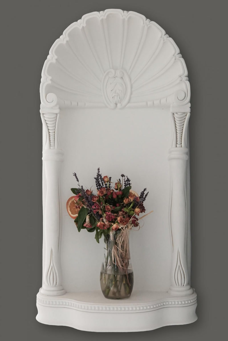 Nișă Decorativă din Poliuretan pentru Perete Model P09 Nișa decorativă din poliuretan reprezintă un element arhitectural monobloc sau prefabricat, destinat creării de adâncime vizuală și spații de expunere pe suprafețe verticale. Modelul P09 este fabricat dintr-un polimer densificat având o densitate de 150-220 kg/m³ (țintă ~160 kg/m³), ceea ce îi conferă o rezistență mecanică ridicată și o stabilitate dimensională excelentă în timp. Cu o rată de absorbție a apei de sub 1%, acest profil nu este afectat de umiditate și rezistă la variații de temperatură cuprinse între -100 °C / +80 °C. Cadrul exterior ornamentat cu coloane canelate și coronament în formă de scoică oferă o structură rigidă ce se poate ancora direct pe perete.

Tehnica de montaj pentru nișa din poliuretan P09 implică pregătirea meticuloasă a suprafeței suport, care trebuie să fie curățată de praf și degresată. Fixarea pe zidărie sau pe plăci de gips-carton se realizează prin aplicarea unui adeziv poliuretanic structural pe spatele piese, combinată cu o fixare mecanică temporară în holsșuruburi. La îmbinările dintre marginea nișei și perete se aplică un mastic acrilic elastic pentru a preveni apariția fisurilor din cauza dilatărilor structurale. Dup uscare, suprafața îmbinărilor se nivelează cu hârtie abrazivă de granulație 220 pentru a obține o trecere invizibilă.

Spre deosebire de nișele tradiționale realizate din gips sau plăci de gips-carton, nișa din poliuretan vine complet turnată din matriță cu toate detaliile sculpturale gata formate. Această caracteristică reduce timpul de executie pe șantier cu peste 70%, eliminând etapele lungi de debitare, ghipsare și șlefuire manuală. Structura celulară închisă a materialului previne apariția mucegaiului și permite o curățare facilă. Produsul este livrat din fabrică acoperit cu o amorsă albă pe bază de apă, pregătită pentru aplicarea straturilor finale de finisaj.

Finisarea nișei P09 necesită aplicarea a cel puțin 2 straturi de vopsea lavabilă acrilică sau pe bază de latex. În aplicația din imagine, albul mat al poliuretanului creează un decupaj geometric clar pe suprafața zugrăvită în nuanță de gri închis, punând în evidență detaliile sculpturale ale nișei. Montajul corect al acestei piese poliuretanice oferă un spațiu de expunere stabil și durabil pentru obiecte decorative, având o durată de viață egală cu cea a structurii pe care este instalată.